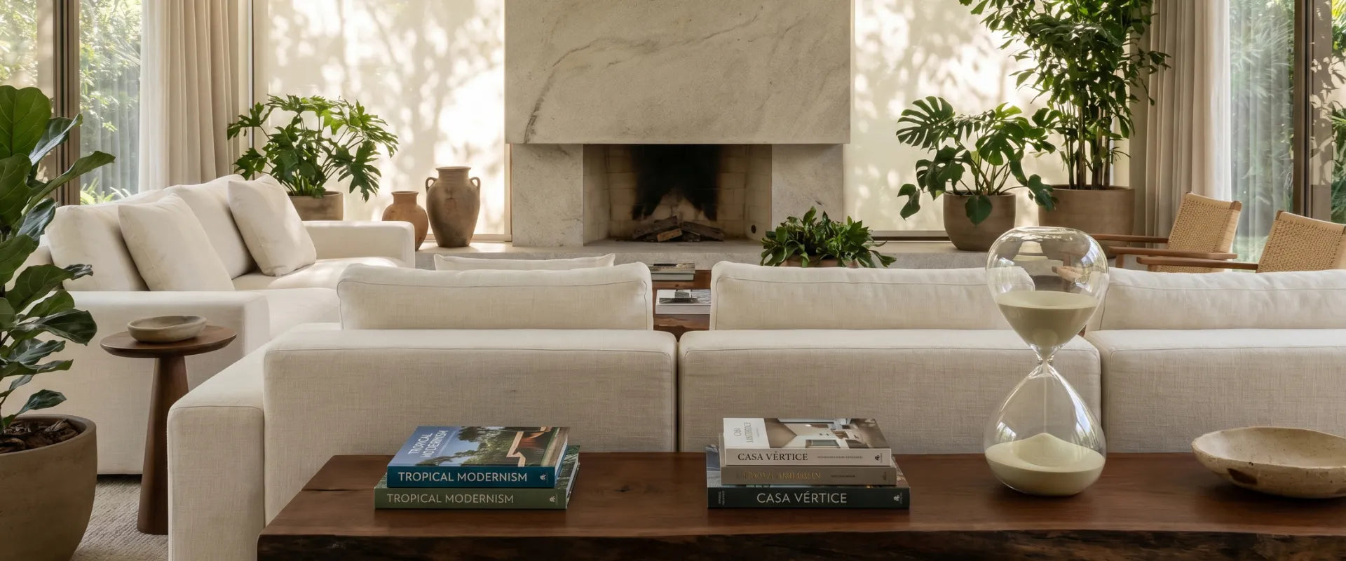 Banner principal da loja CasaVértice Home Decor exibindo uma sala de estar luxuosa e minimalista com arquitetura contemporânea e iluminação natural. No primeiro plano, uma mesa de centro de madeira maciça escura exibe livros de design, um livro com o nome Casa Vértice impresso na capa e uma grande ampulheta decorativa de vidro transparente com areia clara. Ao centro, sofás de linho off-white com almofadas neutras criam simetria em frente a uma lareira monumental de mármore travertino ou pedra natural. Ao fu